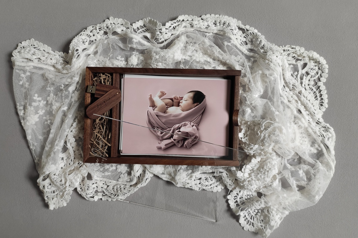 Walnut Wooden Photo Box with acrylic lid displaying a collection of baby photographs on a smooth grey background, accented with delicate white lace fabric